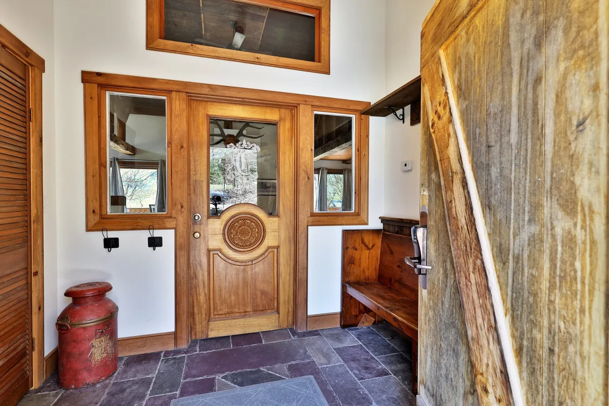 This image features an ornate wooden door with a large window nearby, a wooden bench ideal for sitting while removing boots, and a red metal container that could be used for storing gear. The scene is bright and welcoming, with plenty of natural light.
