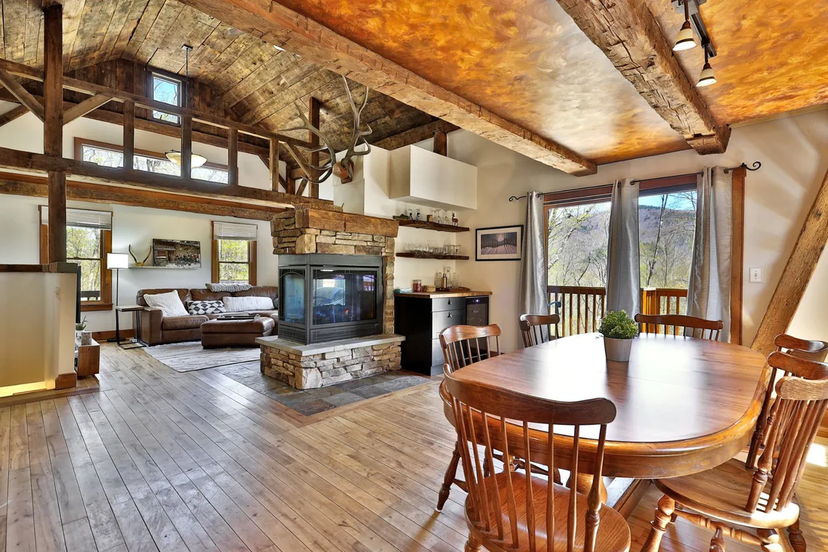 A warmly lit living and dining area featuring a rustic stone fireplace, a wooden dining table, and large windows offering scenic views beyond. The space invites gathering and relaxation, and the windows hint at the surrounding landscape.