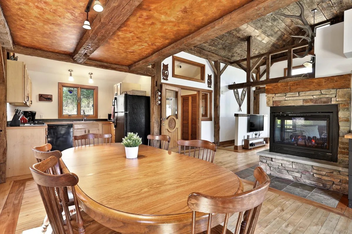 The image features a large wooden dining table set near a stone fireplace, accented by exposed wooden beams overhead. The space feels communal, warm, and ready for memorable meals and celebrations, reinforcing the social aspect of the cabin.
