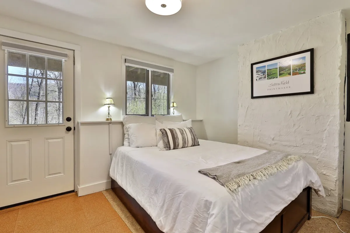 A serene bedroom with a comfortable bed, windows letting in natural light, a door, and artistic touches on the walls. The space feels peaceful and private, suitable for a restful night's sleep.