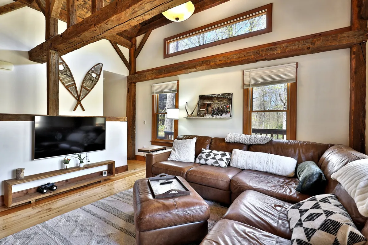The cozy living room is furnished with a large leather sectional sofa, a wall-mounted TV, decorative wooden oars, and expansive windows that fill the space with natural light. The décor nods to adventure and the outdoors while providing a comfortable spot to unwind with family or friends after a day spent exploring.
