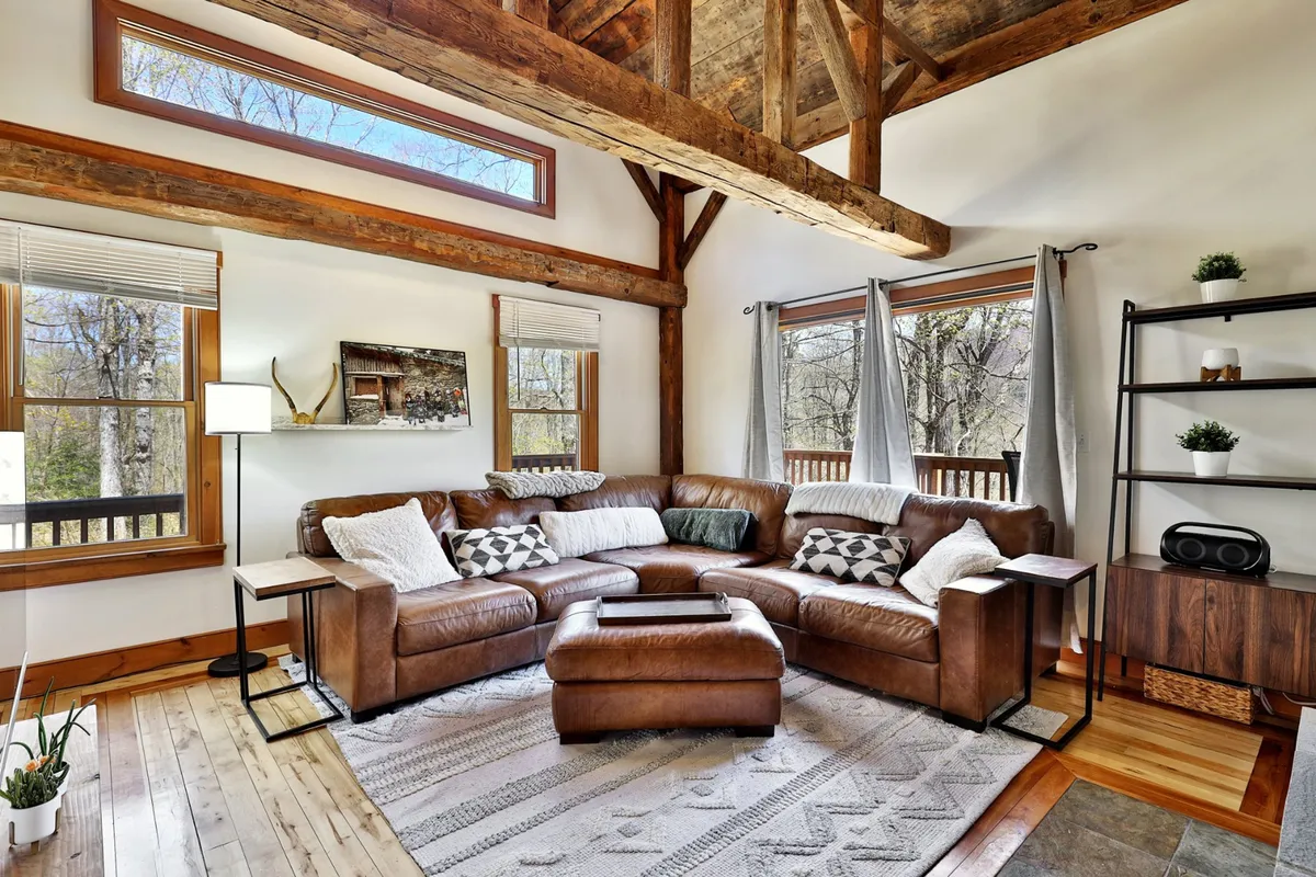 A comfortable seating area with a leather sectional sofa, rustic wooden beams, and expansive windows that flood the room with light. Decorative pillows and a side table create an inviting spot for reading, relaxing, or game nights with the whole family.
