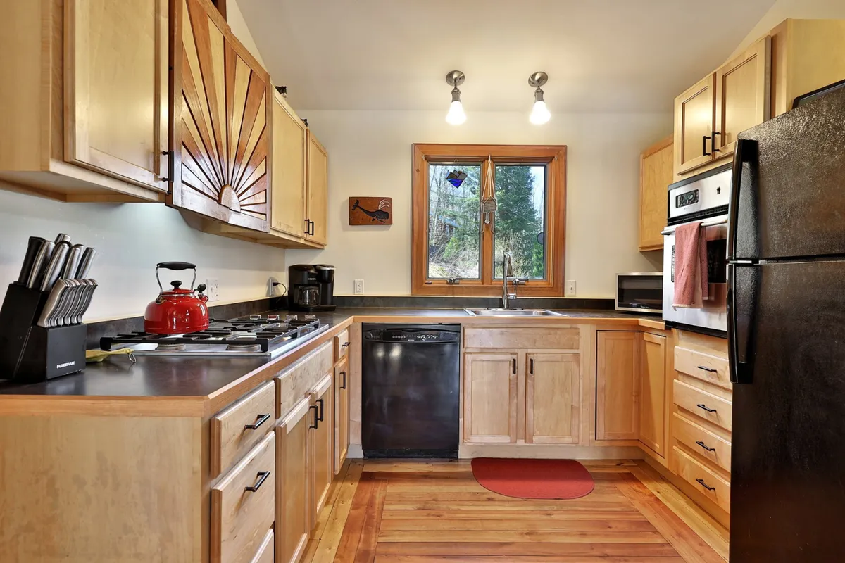 The kitchen features warm wooden cabinetry with a sunburst design, a red kettle, black appliances, and a window offering a view of the surrounding greenery. The space suggests togetherness and a seamless indoor-outdoor connection, hinting at family meals and casual gatherings after time spent exploring.