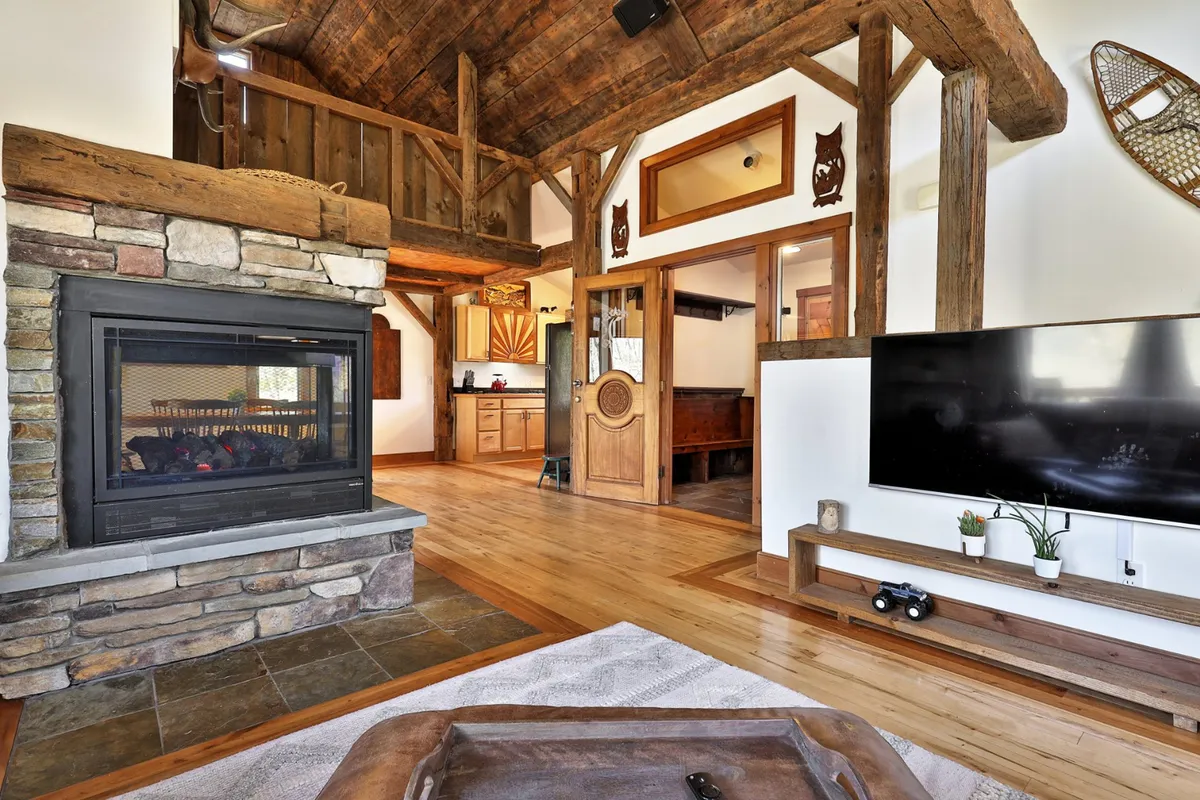 This image showcases a spacious, open-concept living area featuring a glowing fireplace, wooden beams overhead, a large rug, a visible kitchen area, and a television. The scene exudes warmth and togetherness, with plenty of seating for a family to gather, play games, or enjoy a cozy movie night.