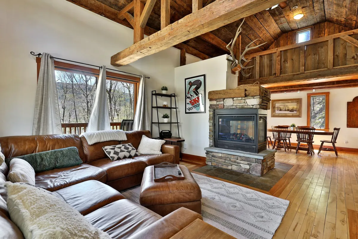 This image showcases an open-concept living space featuring a leather sectional sofa, a warm fireplace, expansive windows with scenic views, and the adjacent dining area. The setting is bathed in natural light, creating a welcoming environment ideal for gatherings, relaxation, and enjoying the outdoors from the comfort of the interior.
